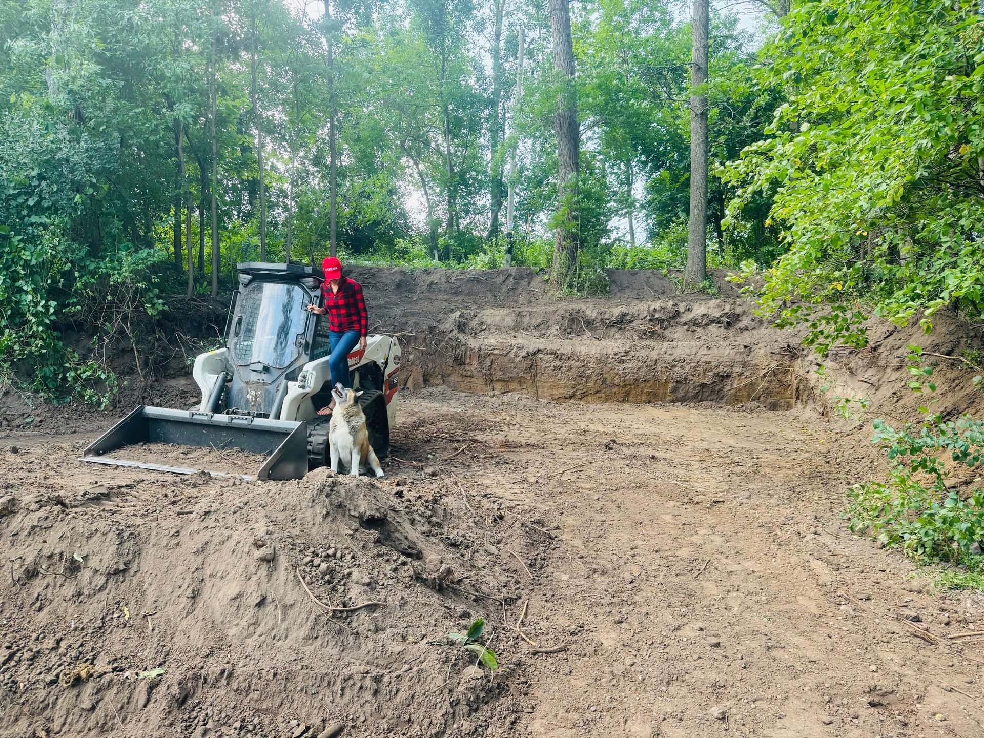 Person operating a tractor in a wooded area; dog stands nearby. The ground is dirt, with tiered sections.
