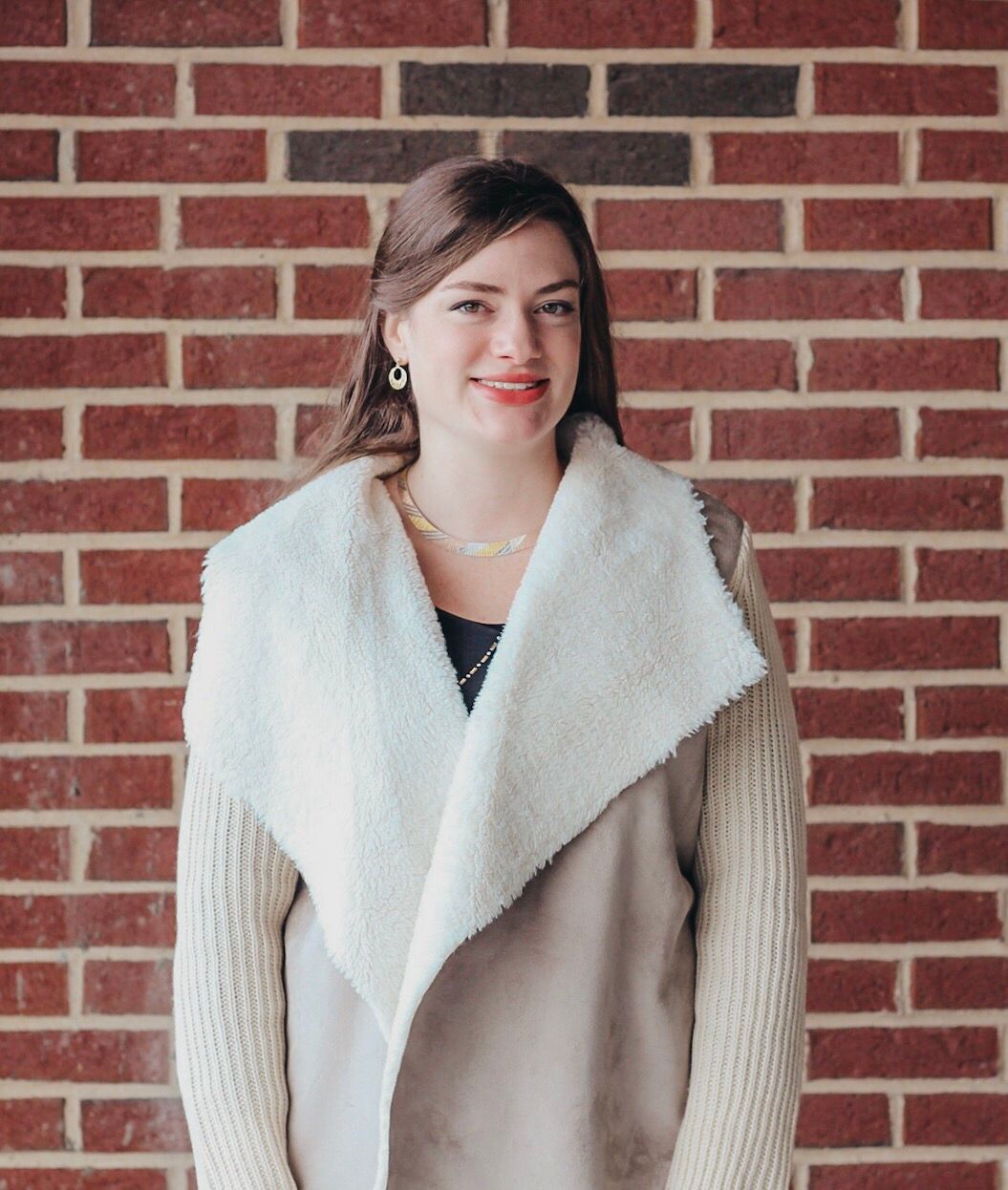 A woman in a white coat is standing in front of a brick wall.