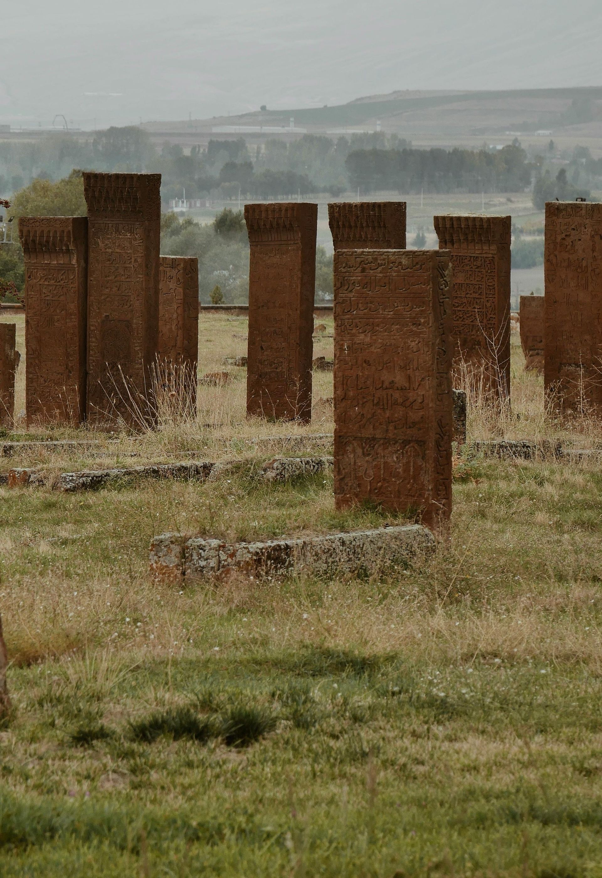 Tall, rectangular brick headstones in a grassy field with a hazy, distant landscape backdrop.