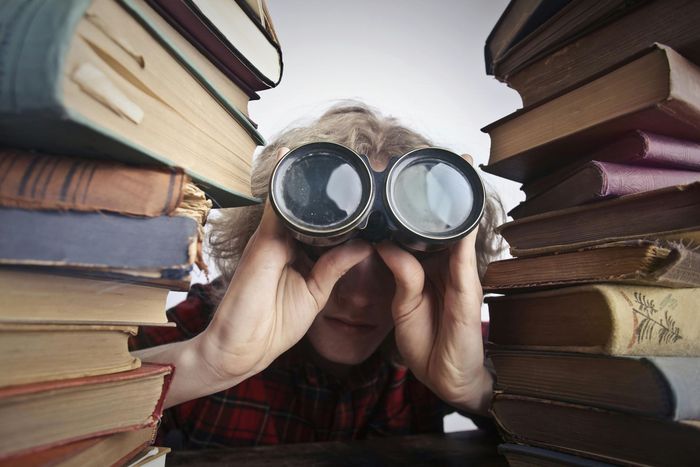 A person is looking through binoculars in front of a pile of books.