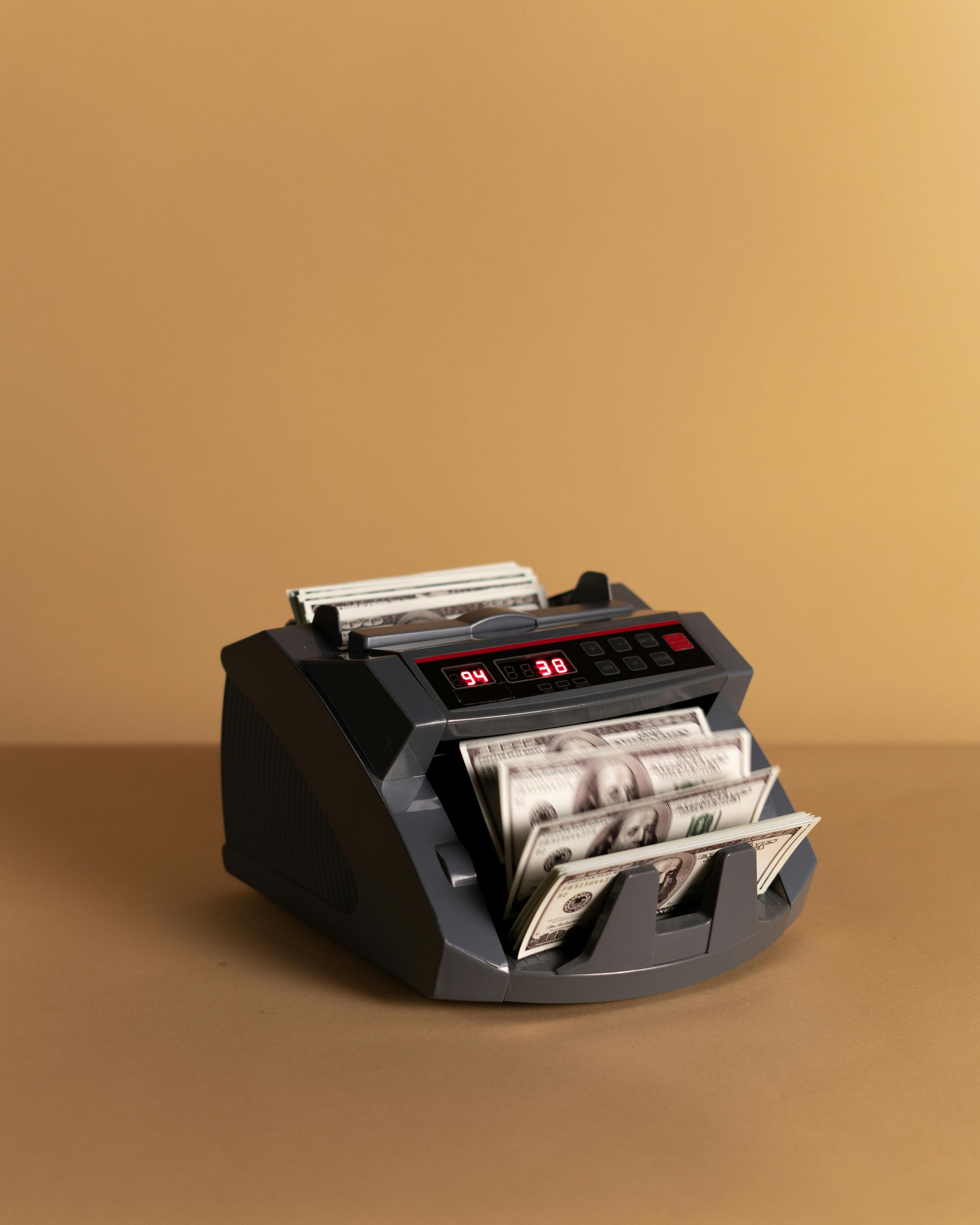 Money counting machine, gray, with bills stacked in input and output trays, brown background.