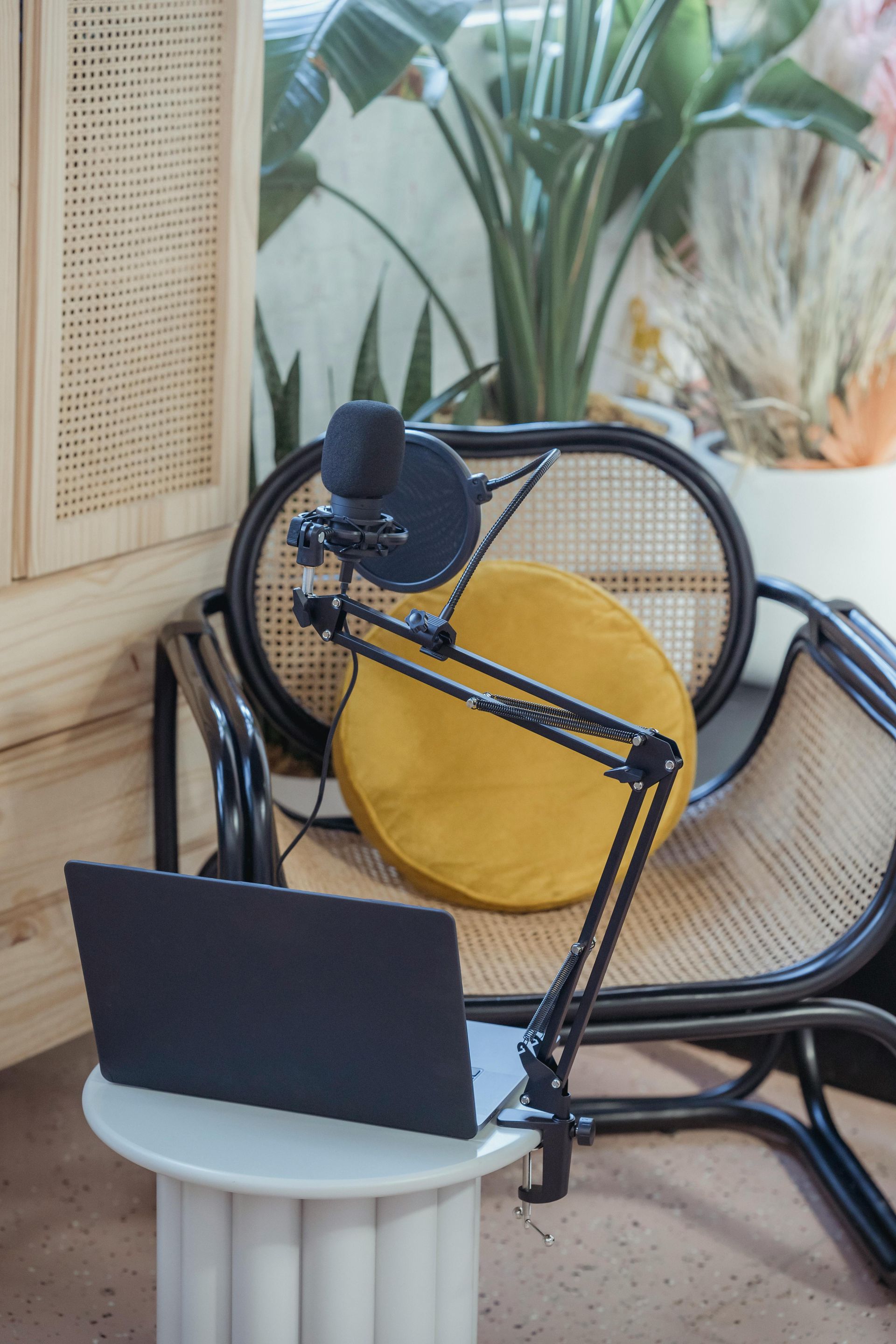 Black microphone and laptop on a round white table next to a wicker chair with a yellow pillow.