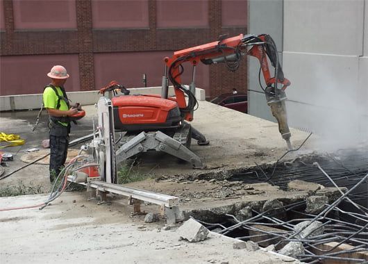 A man is standing next to a robotic concrete demolition machine that is breaking concrete.