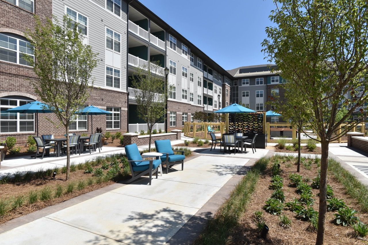 Outdoor communal courtyard with seating, blue umbrellas, and landscaped walkways beside a modern apartment building.