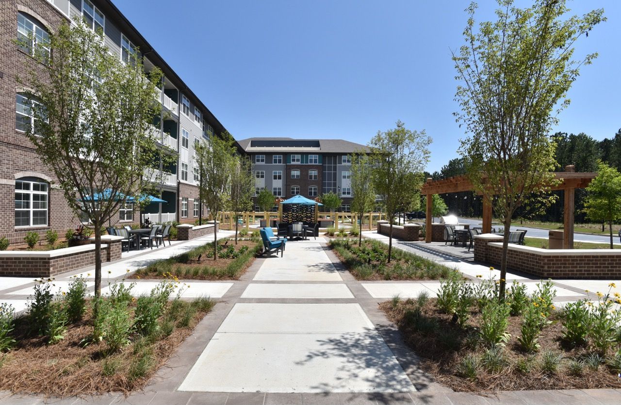 Outdoor communal courtyard with tables, chairs, trees, and a pergola between apartment buildings.