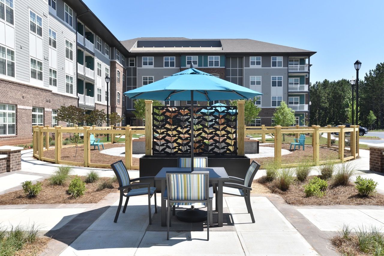 Outdoor communal seating area with table, chairs, and a blue umbrella in a landscaped courtyard.