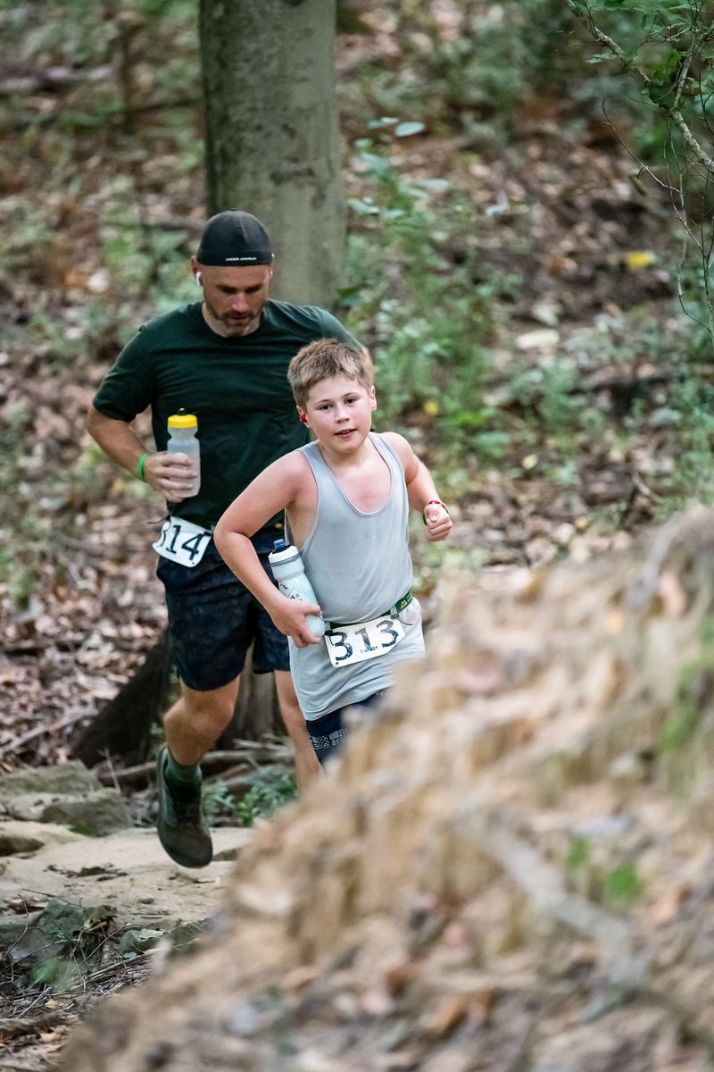 A man and a boy are running down a trail in the woods.