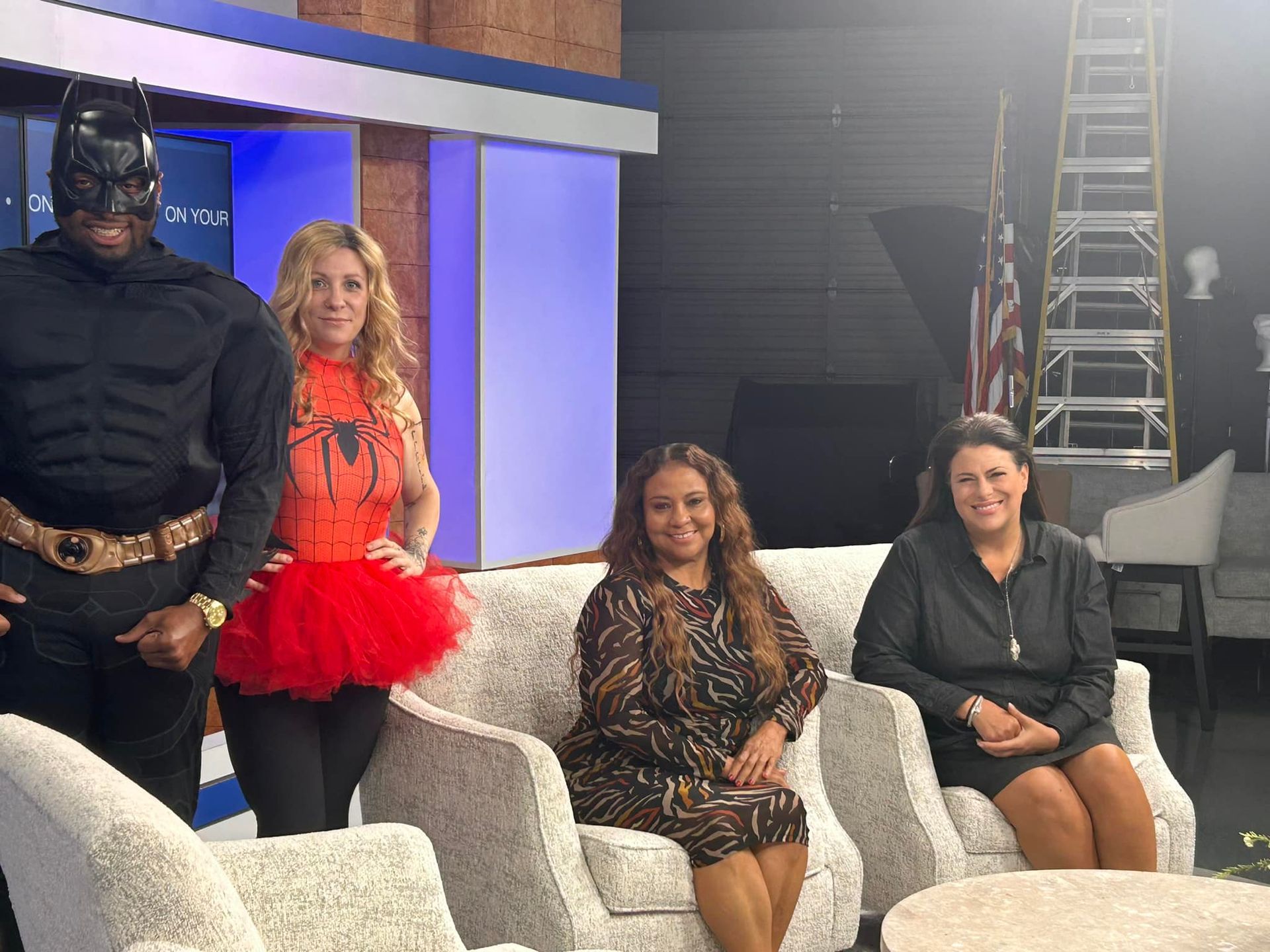 Four people pose on white chairs in a studio set, including a Batman-costumed figure and a woman in a red tutu.