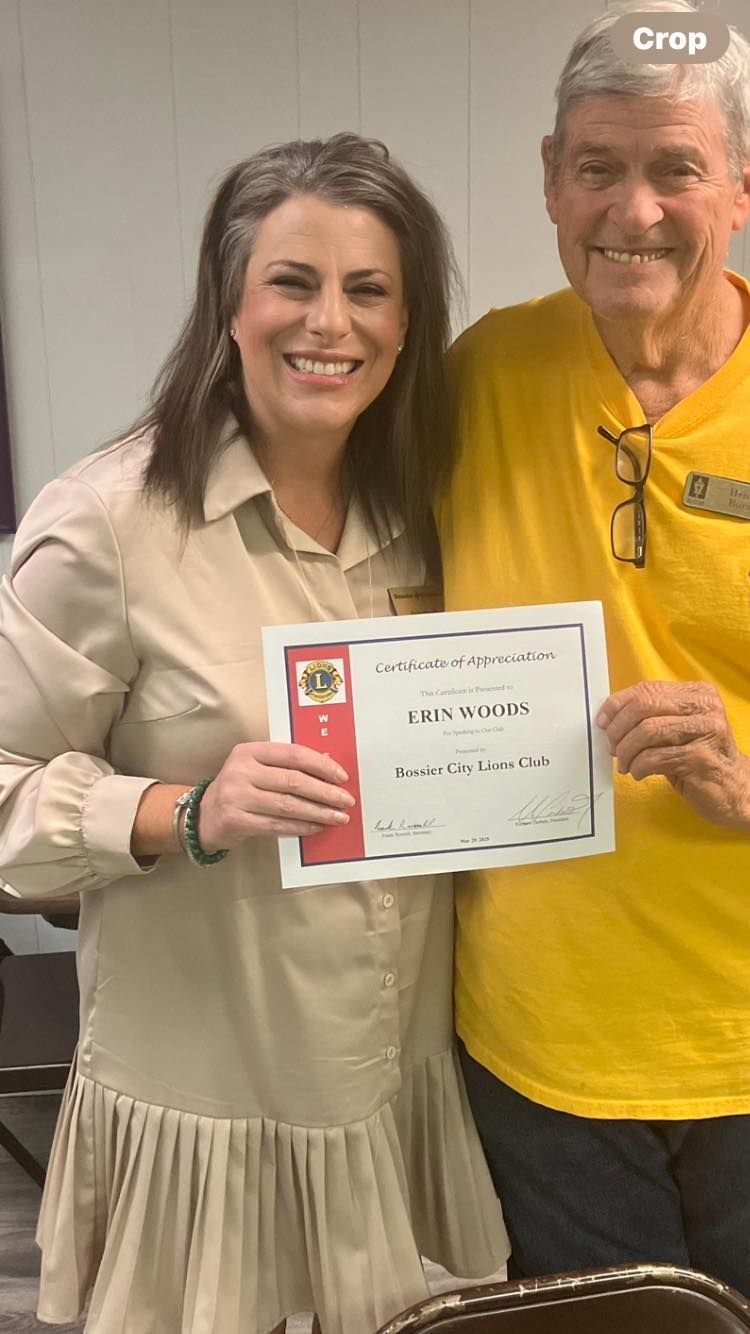 Two people smiling, holding a certificate indoors; one wears beige, the other a yellow shirt.