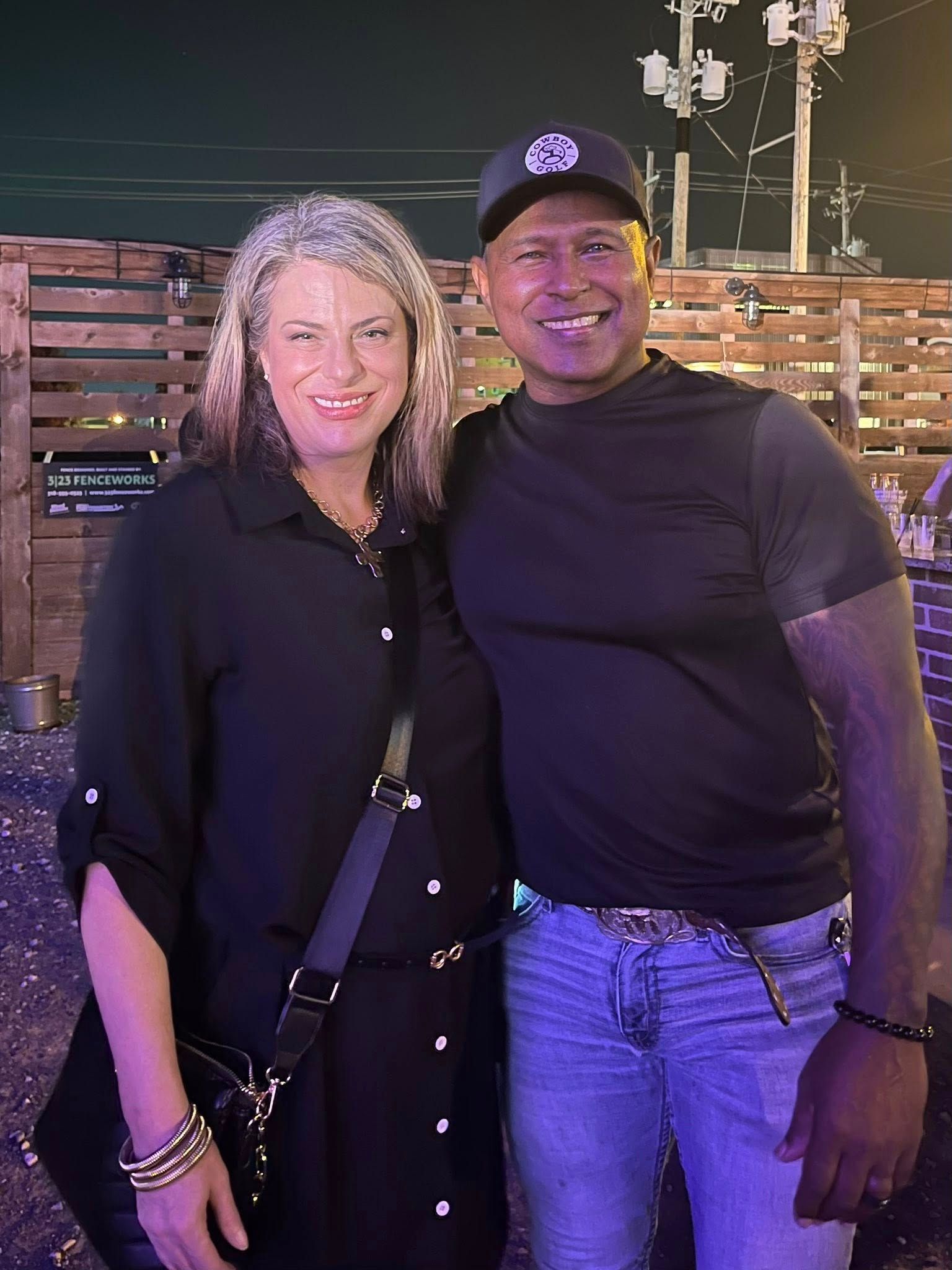 Two people smiling and posing together at night, wearing black tops under purple lighting.