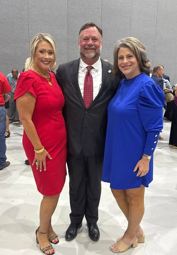 Three people posing at an indoor event, with one in a red dress, one in a suit, and one in a blue dress.