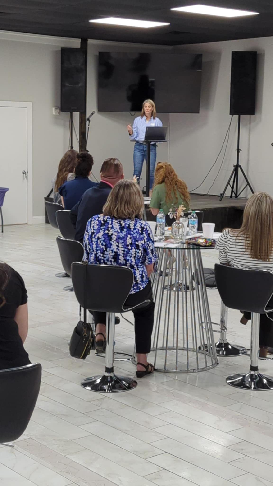 Speaker at a podium addressing an audience in a room with large speakers and TV screens.