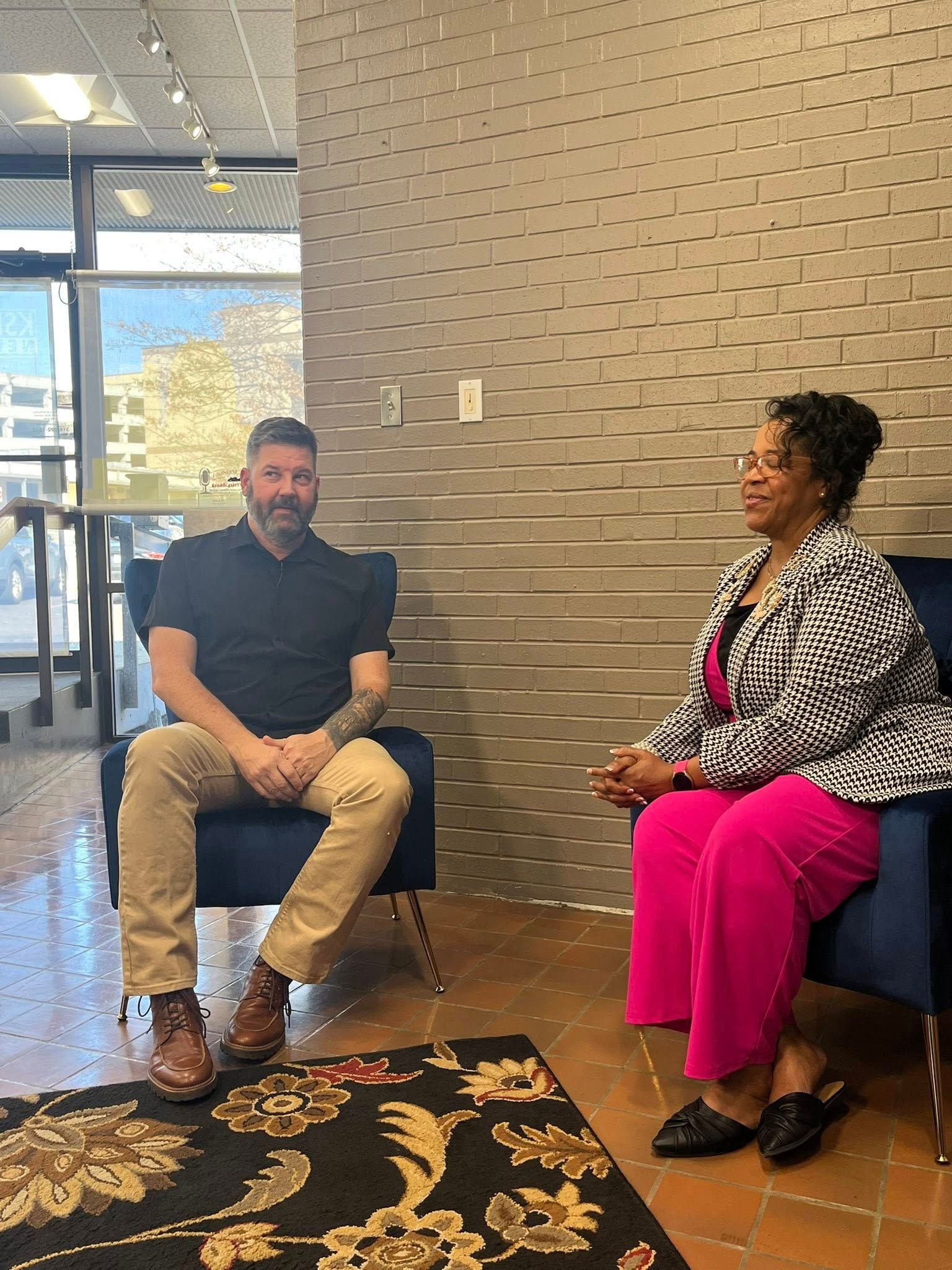 Two people seated in conversation inside a modern lobby with a textured wall and patterned rug.
