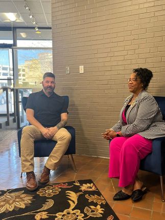 Two people seated in conversation inside a modern lobby with a textured wall and patterned rug.