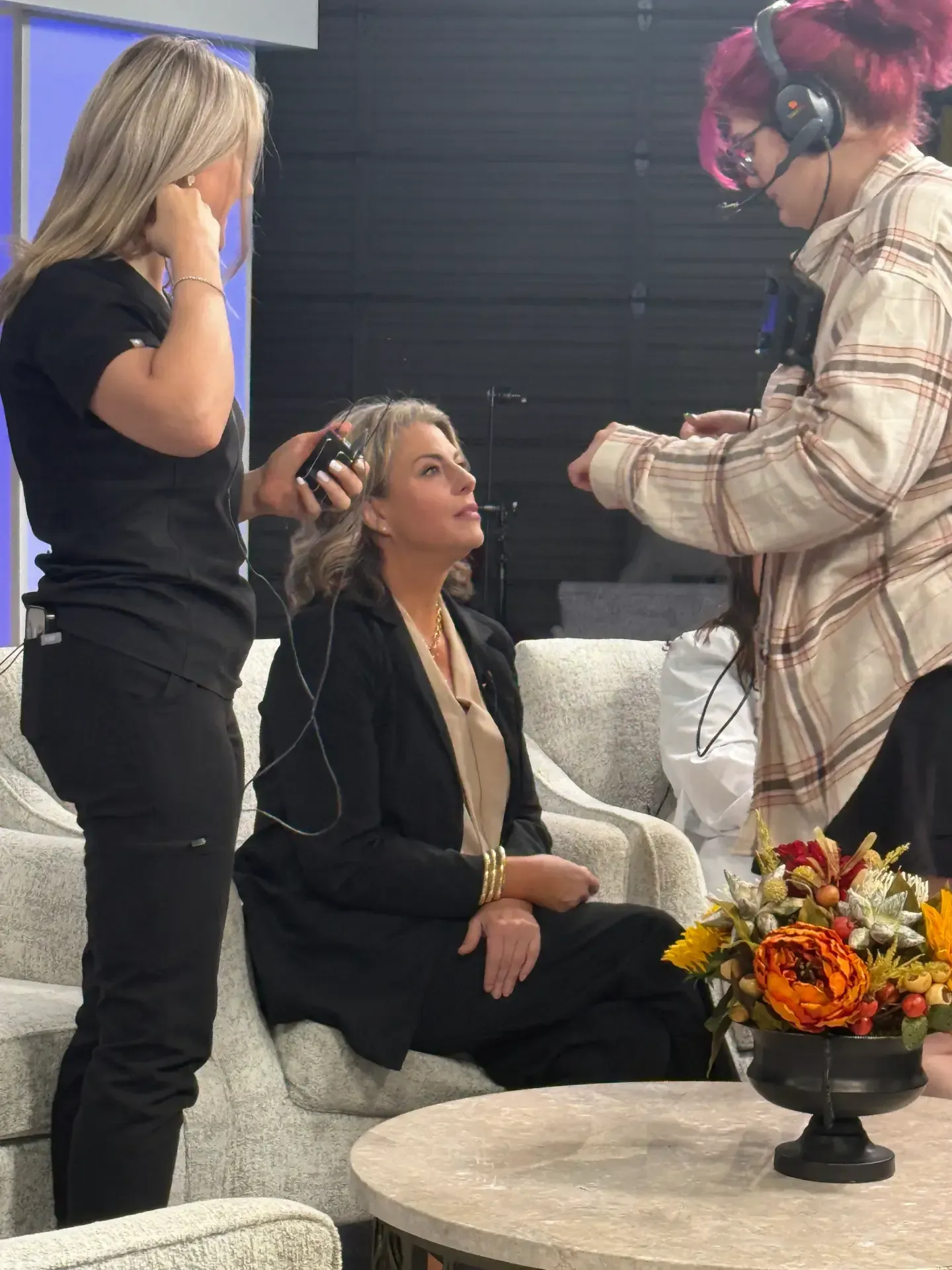 Two people with headsets assist a seated woman in a studio lounge area.