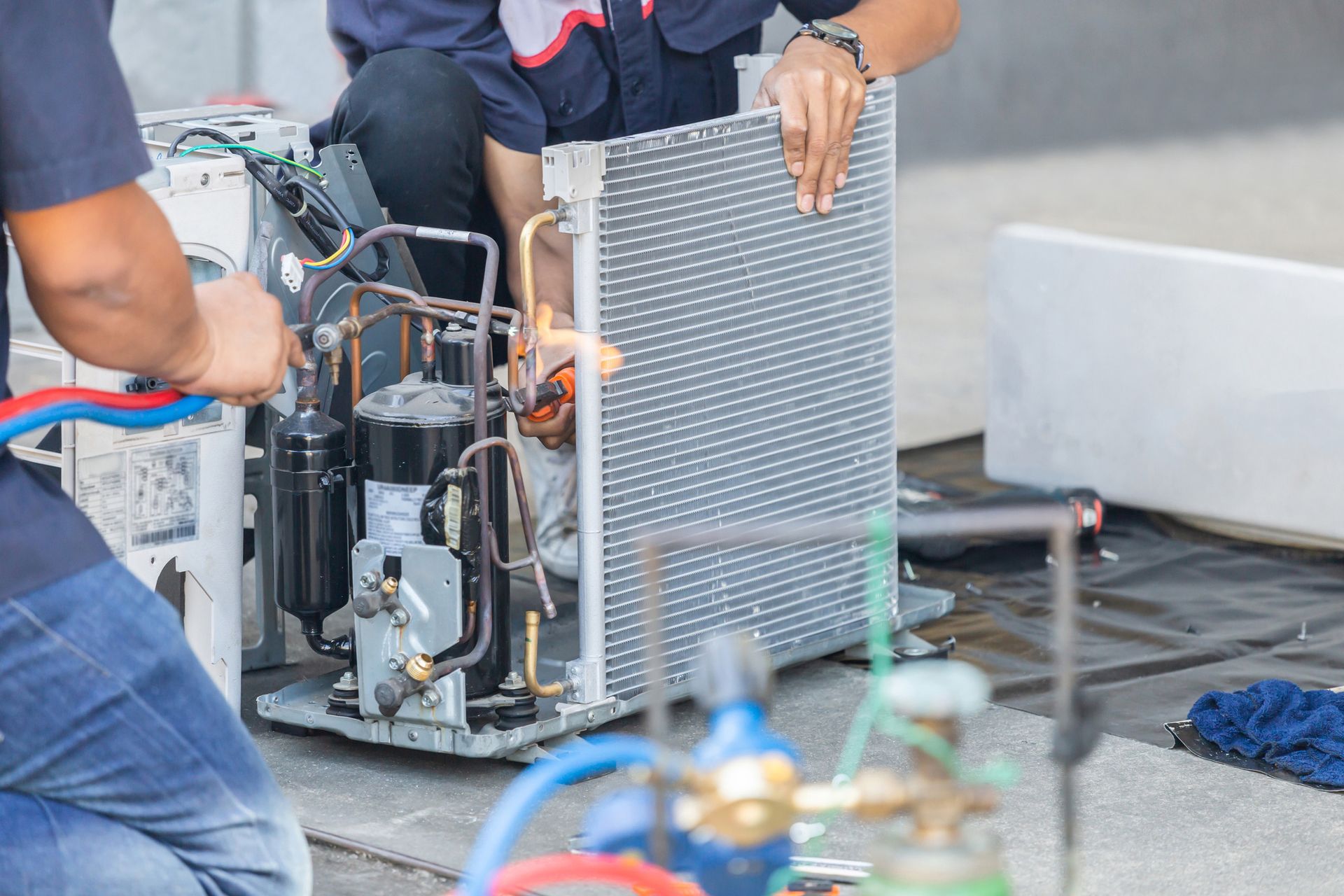 Two HVAC technicians working on an air conditioner, using tools and a heat source outdoors.