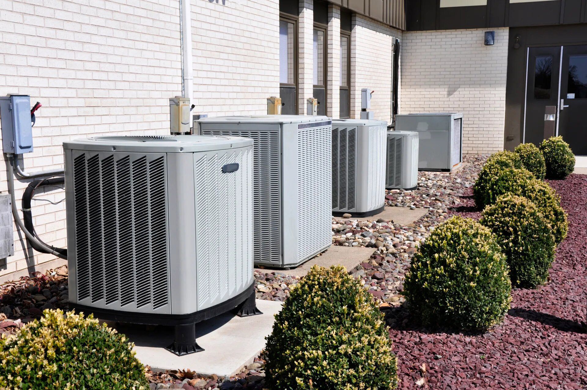 Air conditioning units lined up outside a brick building with landscaping.