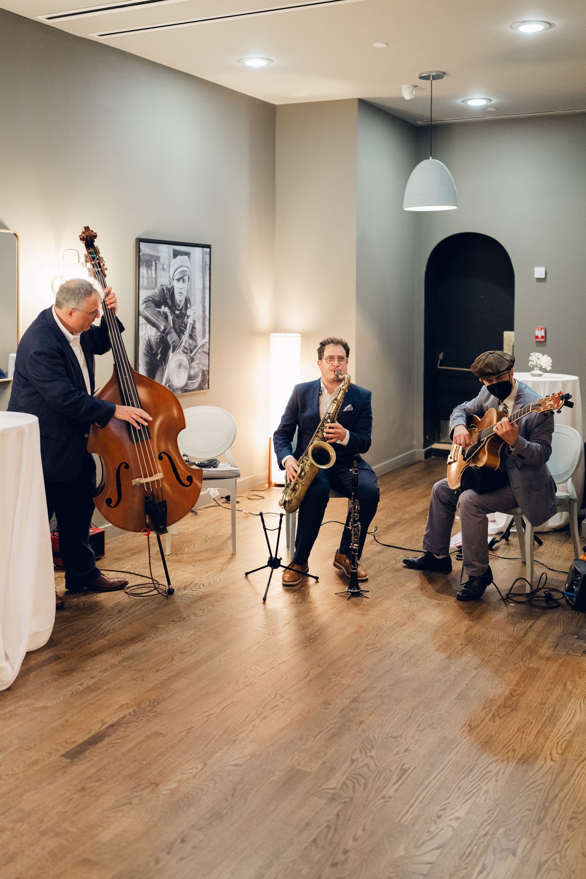 Jazz trio: bassist, saxophonist, and guitarist perform in a room with a wood floor and neutral walls.