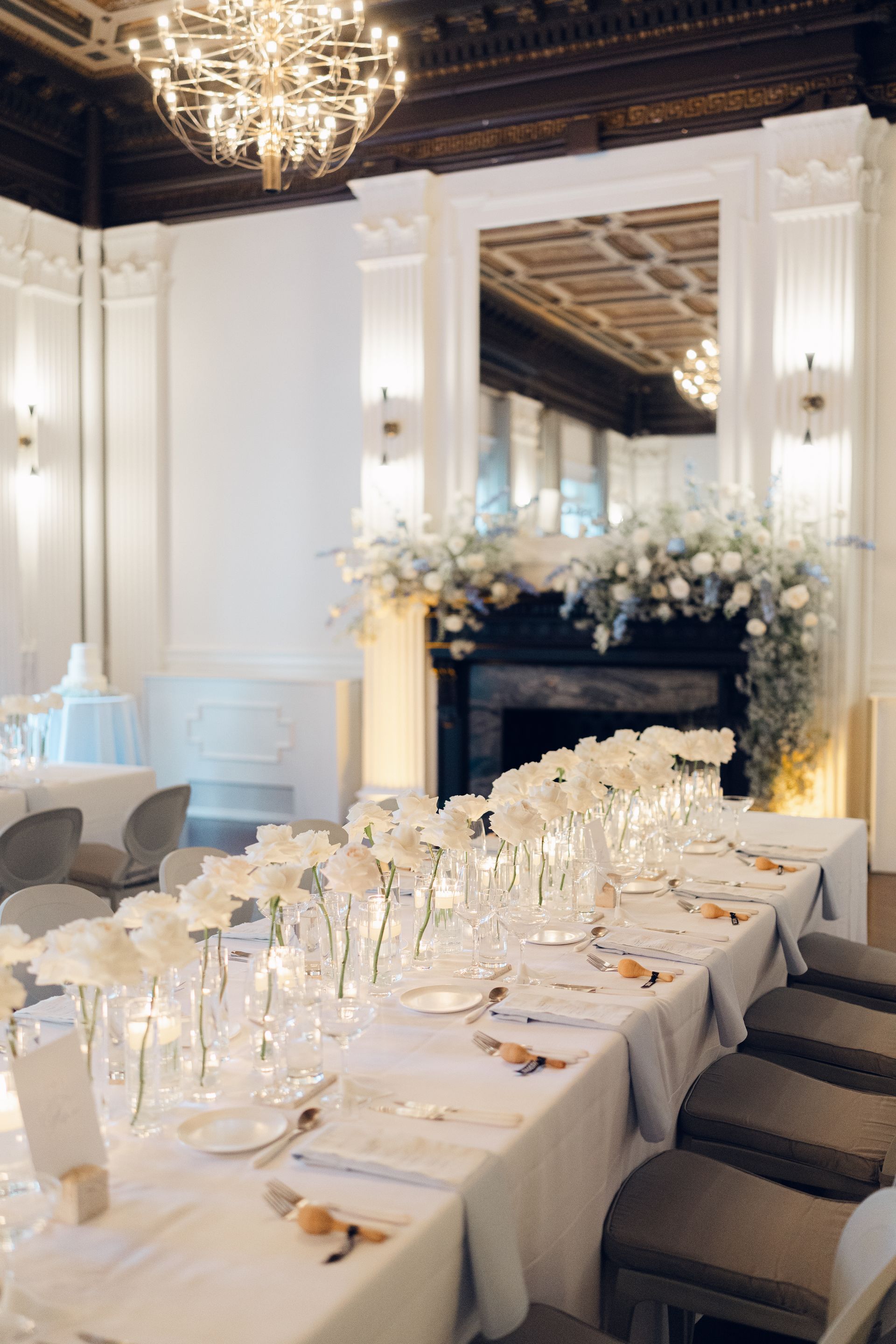 Elegant reception table set with white flowers and glassware, fireplace backdrop, chandelier, and gray chairs.