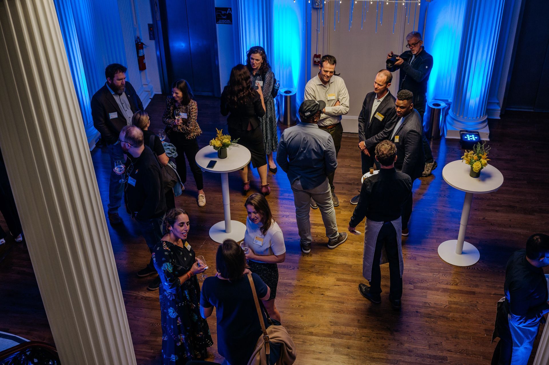 People socializing in a room with blue lighting, round tables, and white columns.