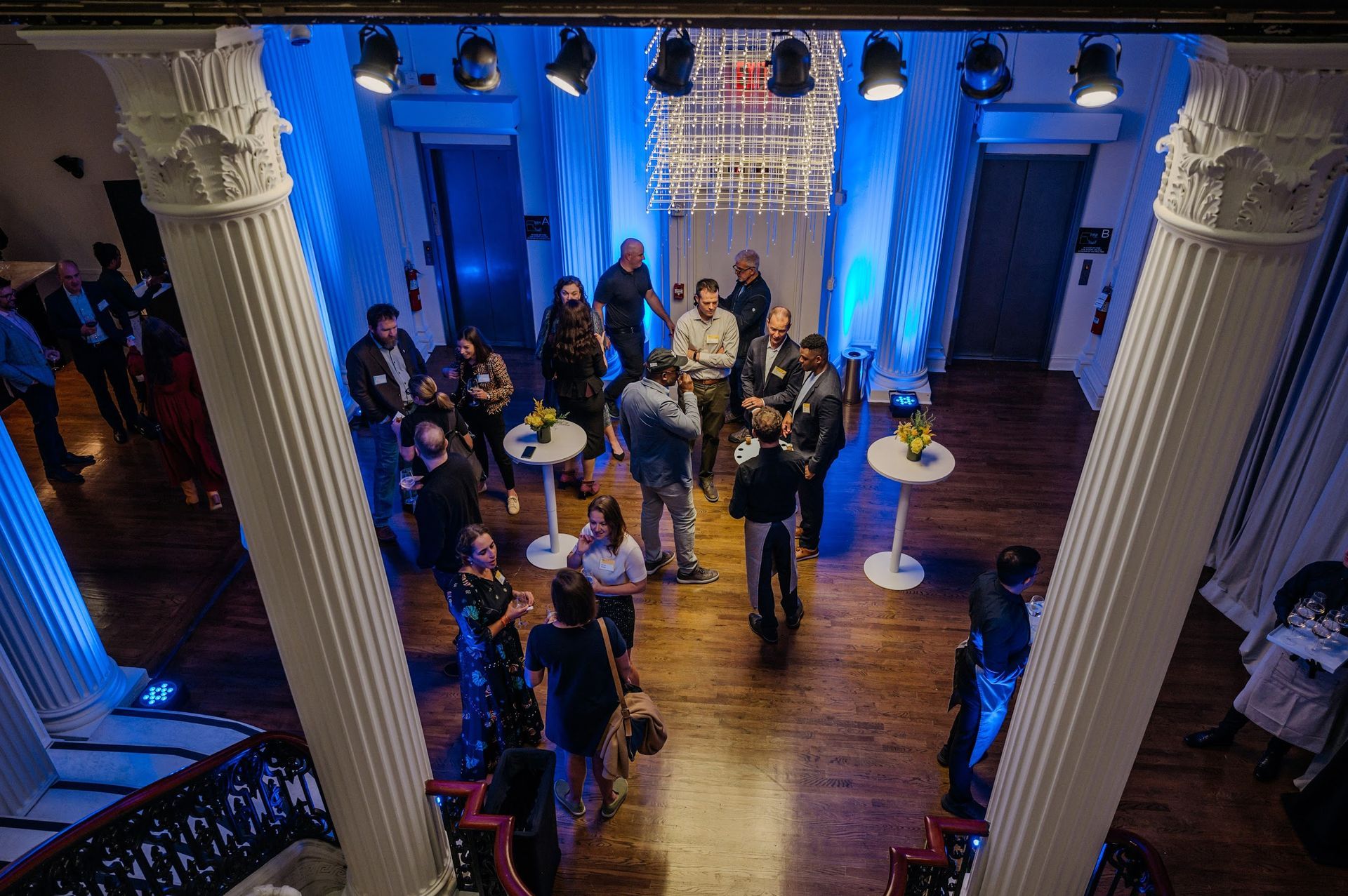 A group of people are standing around tables in a room with columns.