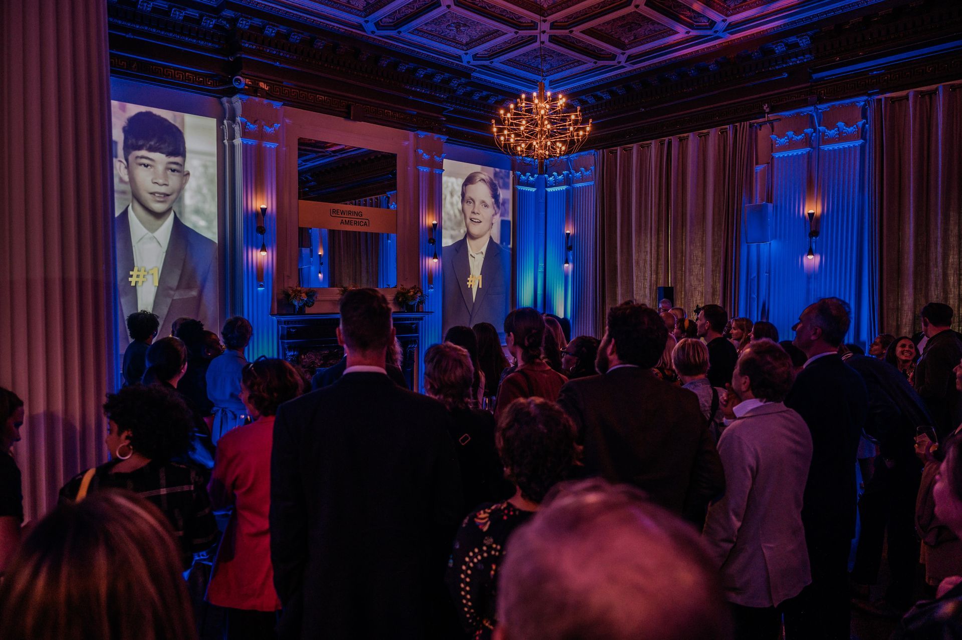 People at an event with projected portraits. Room with blue and purple lighting, chandelier, and curtains.
