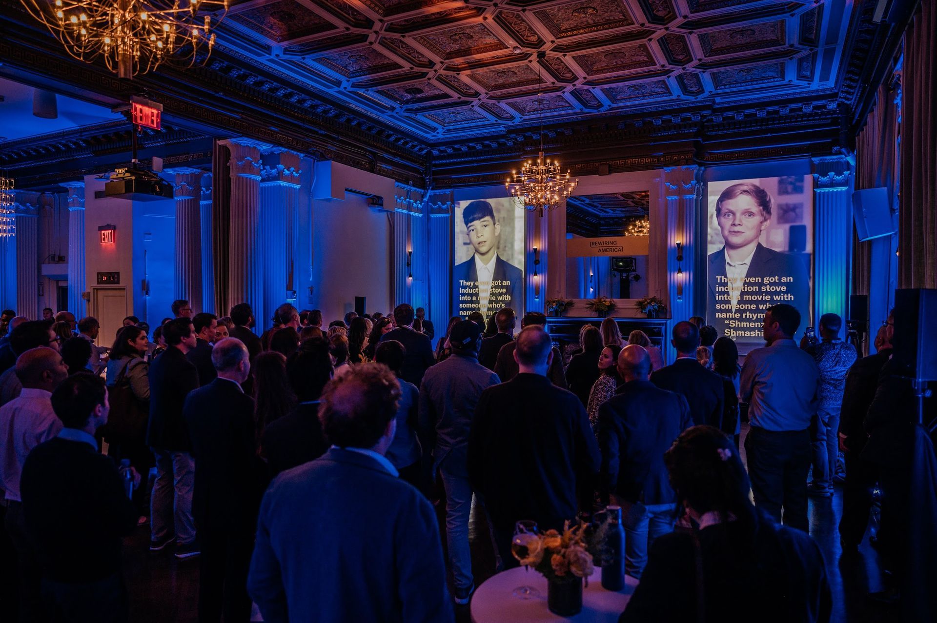 A large group of people are standing in a room with a projector screen projected on the wall.