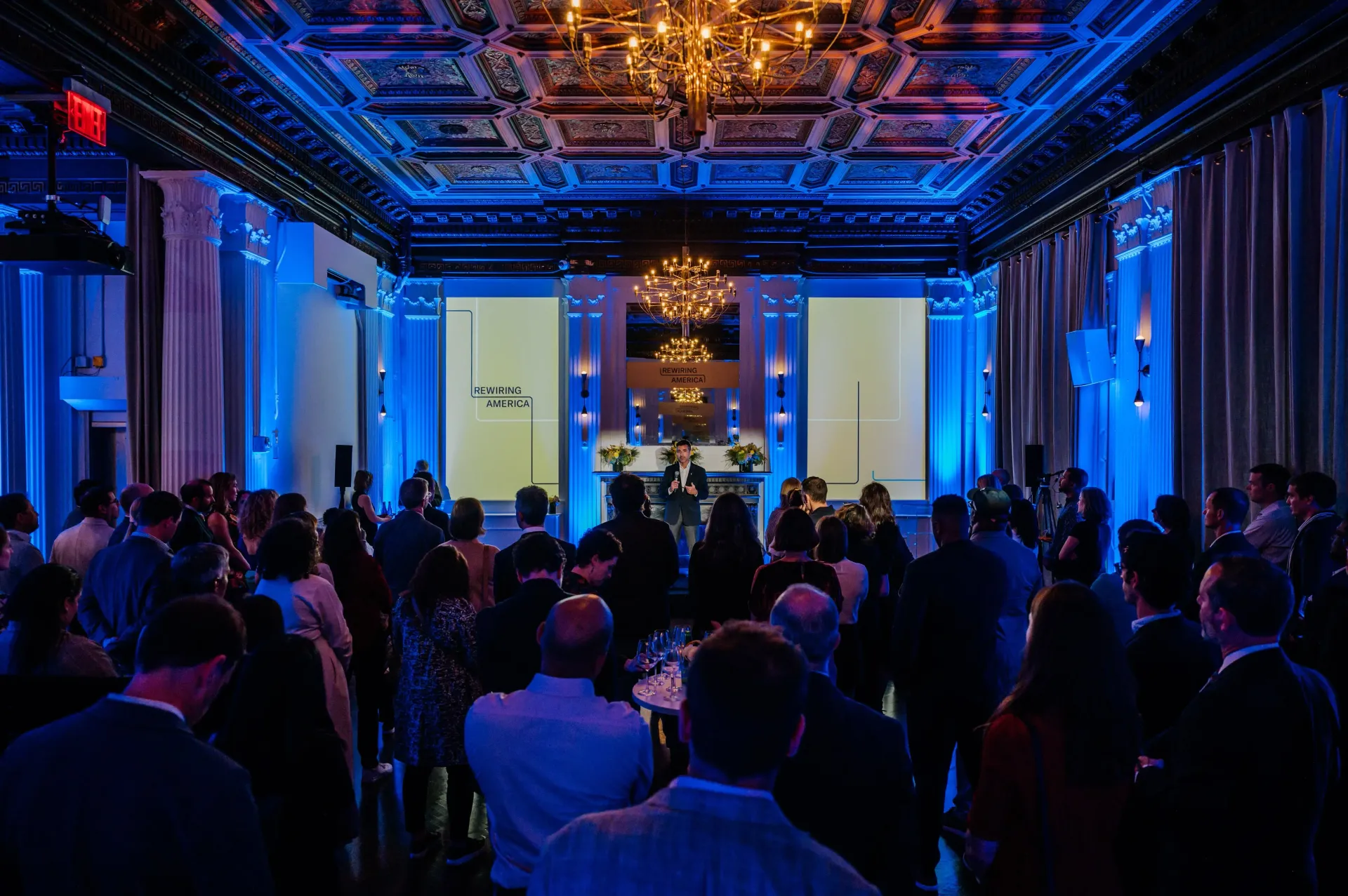 People gathered in a dimly lit event space with blue lighting and a stage. A chandelier hangs from the ceiling.