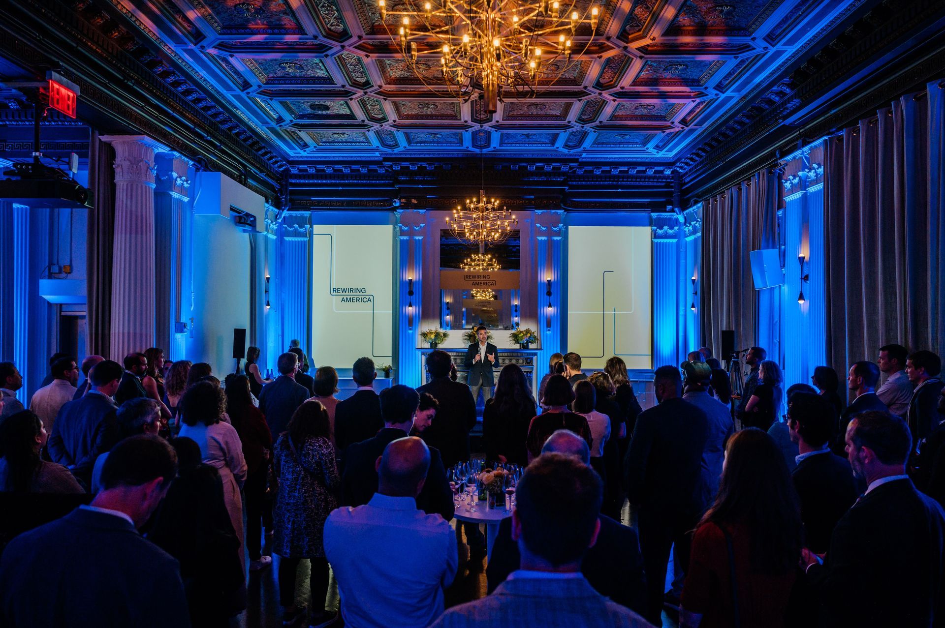 People attending a presentation in a grand hall with blue lighting and chandeliers.