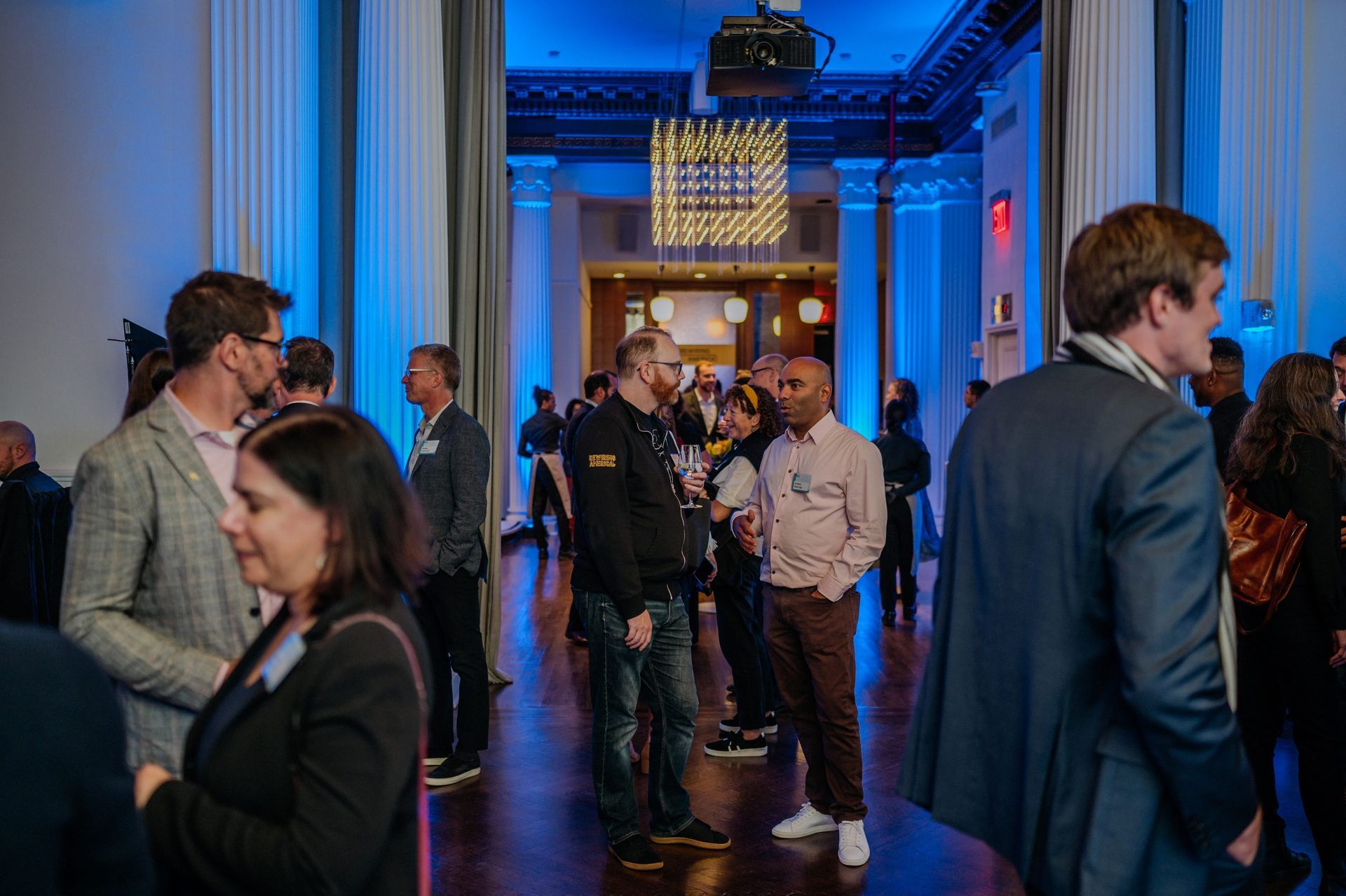 People mingling in a long room with blue lighting; event setting with columns and decorative light fixtures.