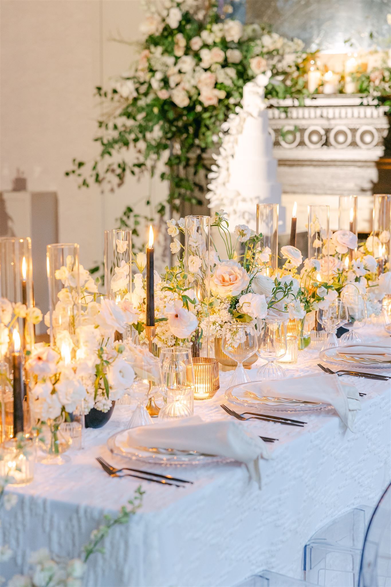 Elegant dining room with long tables set for a formal event. White linens, silver chairs, and crystal chandeliers.