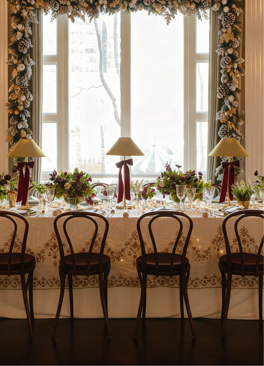 Elegant dining table set for a holiday meal, with lamps, floral arrangements, and four chairs in front of a window.