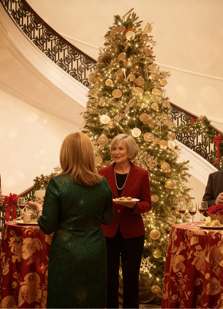 Woman in green dress talks to woman in red blazer holding cookies near Christmas tree.