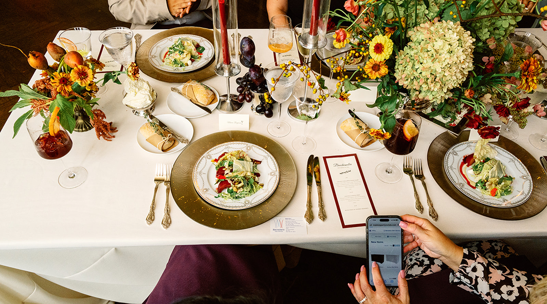 Table setting with gold chargers, floral arrangements, food, and someone taking a photo with a phone.