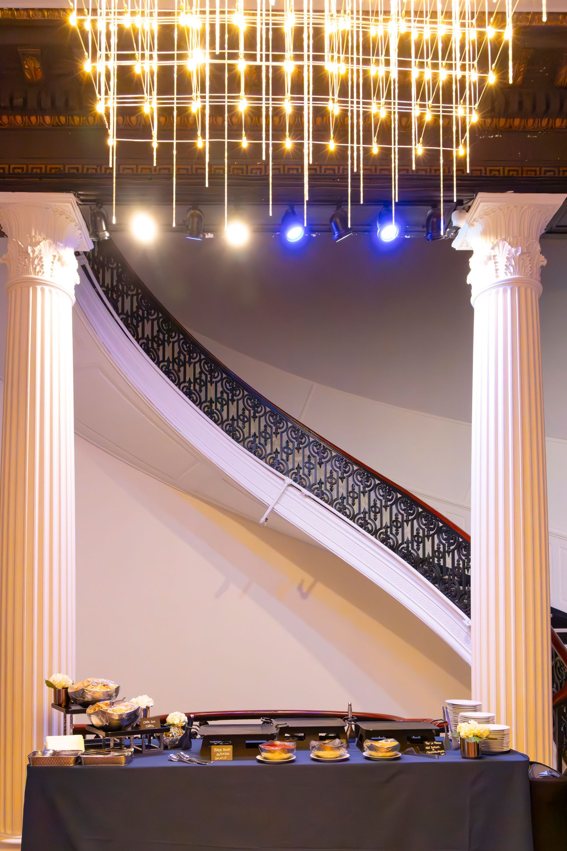 Buffet table set below a grand staircase, beneath a decorative chandelier and columns, with spotlights.