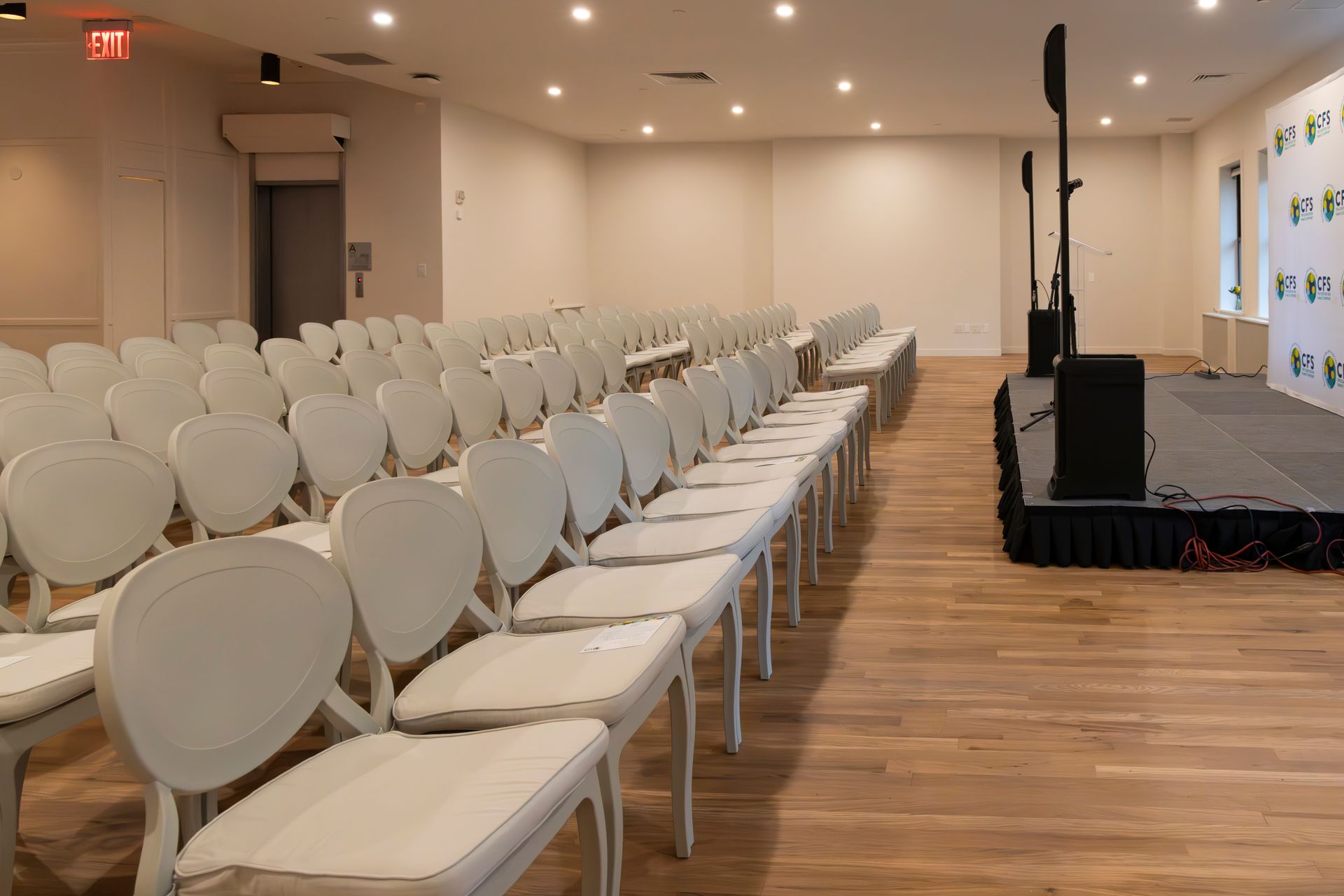 Empty event space with rows of white chairs facing a stage with sound equipment. Wooden floor, cream walls.