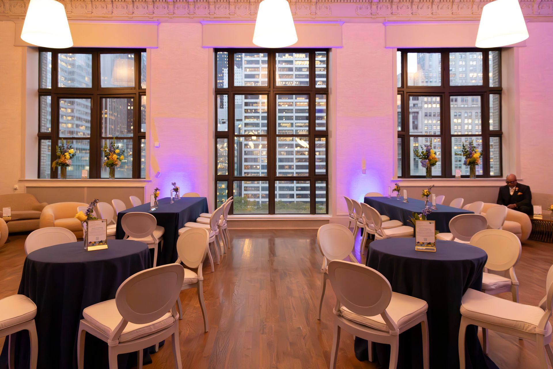 Event space with round and rectangular tables, white chairs, and large windows with city views.