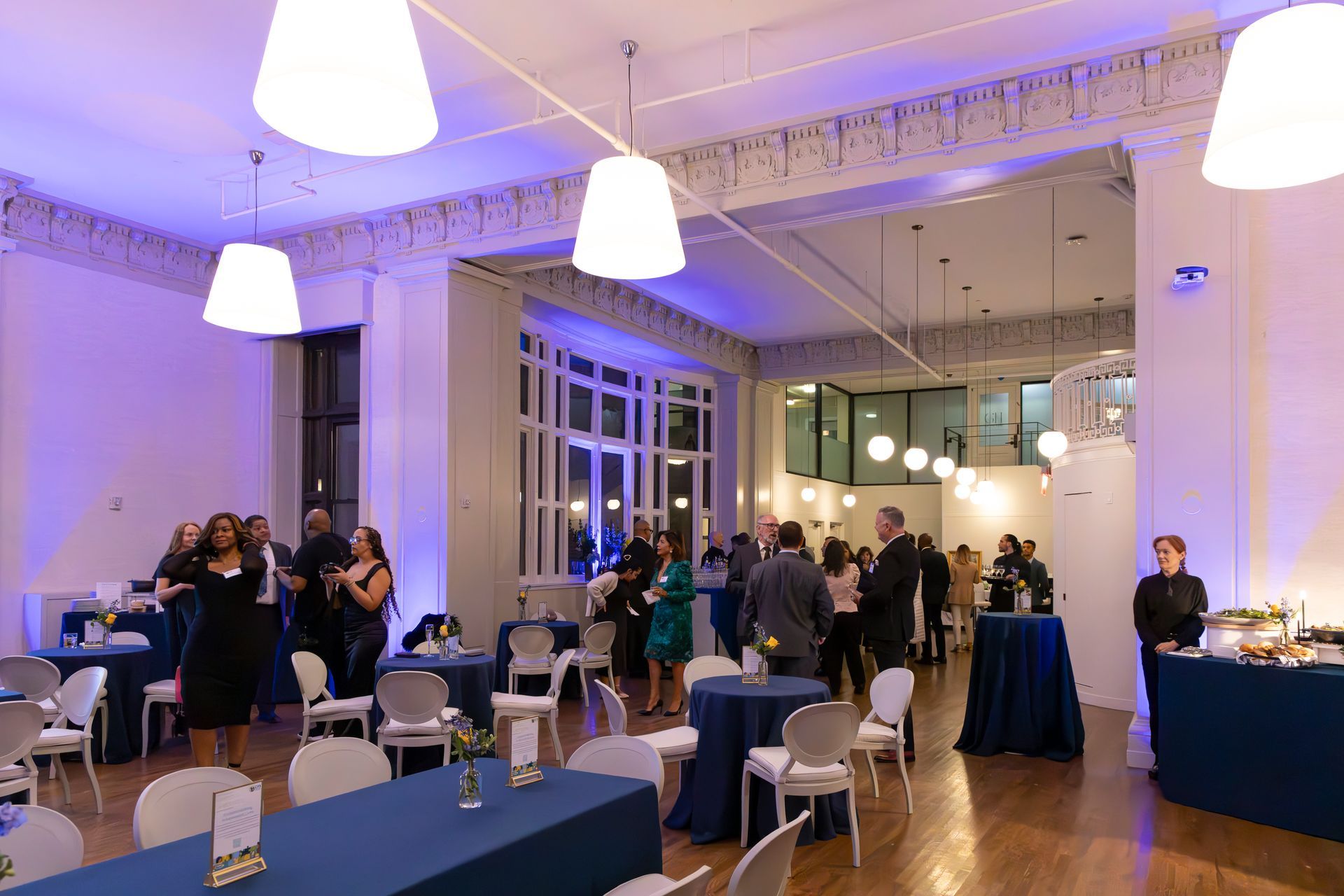 Event in a room with blue lighting, tables, and people socializing.