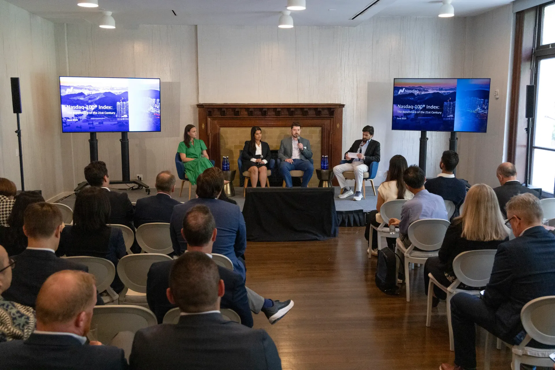 Panel discussion in a room with a seated audience, two screens displaying images, and five panelists at a table.
