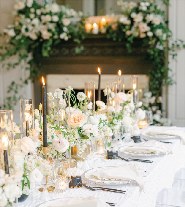 Elegant wedding table setting with white flowers, candles, and a decorated fireplace.