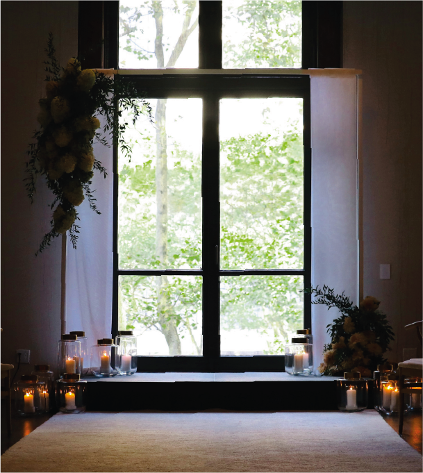 Wedding ceremony setup: large window backdrop, floral arrangements, candles, and a white aisle runner.