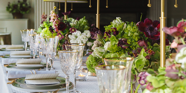 Table setting with gold chargers, floral arrangements, food, and someone taking a photo with a phone.