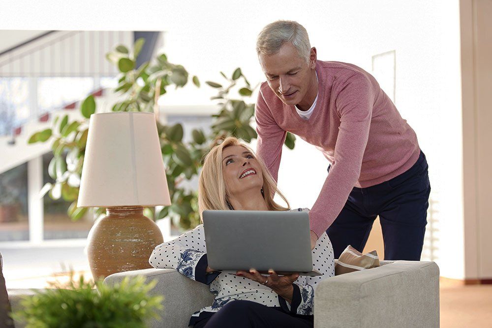 A person sitting in an armchair looks up at another person standing behind them while they both use a laptop.