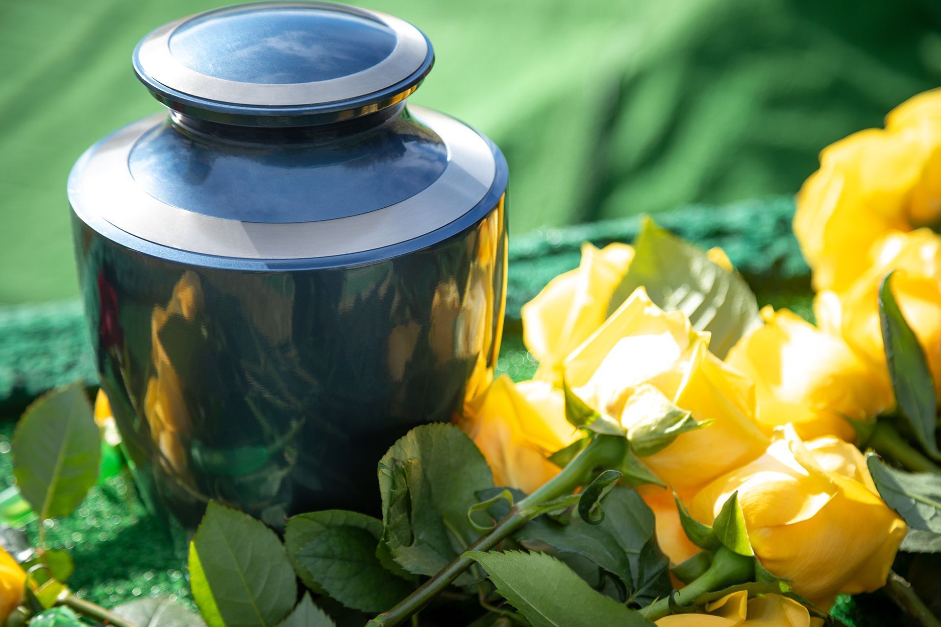 A polished blue cremation urn rests next to a bouquet of yellow roses on a green surface.