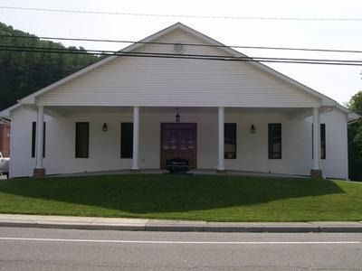 A white, single-story building with a front porch, columns, and central double doors, situated on a grassy lot.
