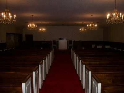 A dimly lit church sanctuary with rows of wooden pews facing a central altar under four hanging crystal chandeliers.