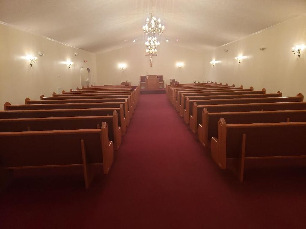 A church interior with rows of wooden pews lining a red carpeted aisle leading to a podium beneath a hanging chandelier.