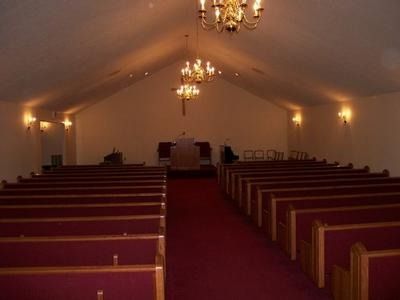 Interior of a chapel with rows of red-cushioned wooden pews facing a raised pulpit under warm pendant lights.
