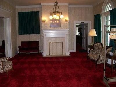 A room with red carpet, a central fireplace, a patterned armchair, a dark sofa, and hanging green curtains.