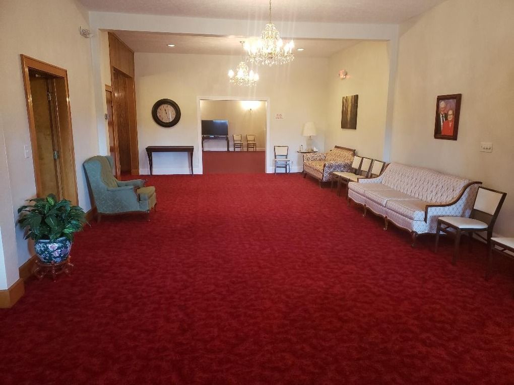 A spacious room with red carpeting, a teal armchair, patterned couches, wooden side tables, and two chandeliers.